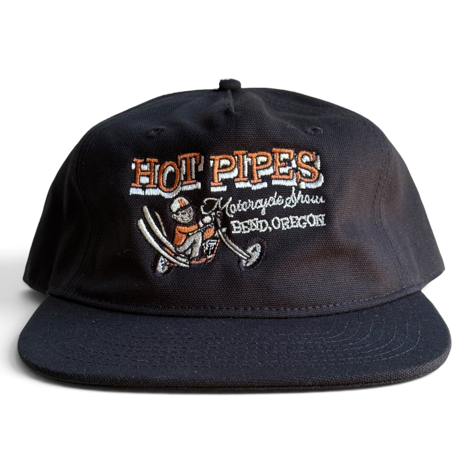 A vintage inspired black flat-brim canvas cap featuring embroidered text and a graphic on the front. The embroidery reads 'HOT PIPES Motorcycle Show BEND, OREGON' with an illustration of a person on a vintage motorcyle. 